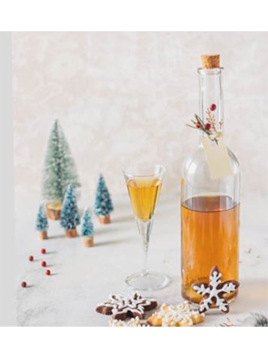Selbstgemachter Bratapfellikör in einer Glasflasche mit Korken, serviert im Likörglas neben winterlichen Plätzchen und Miniatur-Tannenbäumen