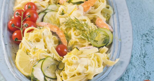 Bild von einem tiefen Teller, in welchem Bandnudeln in heller Sauce, Gurken, Tomaten, Dill und Lachs gemischt angerichtet sind.